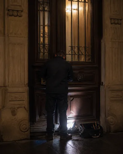 Cerrajero de espalda con linterna de trabajo frente a un portal de vivienda, sin rostro visible.