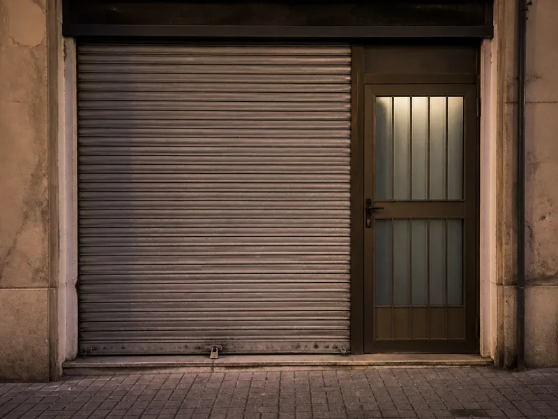 Persiana metálica y puerta peatonal de un comercio, acera vacía, sin rótulos.